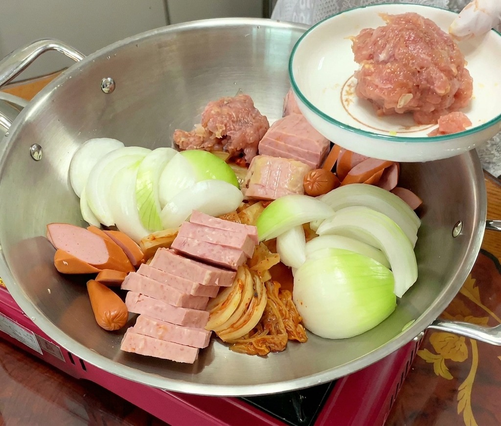 Porcja różowej, mielonej wieprzowiny jest dodawana do garnka wypełnionego już ułożonym kimchi, mielonką, parówkami i cebulą.