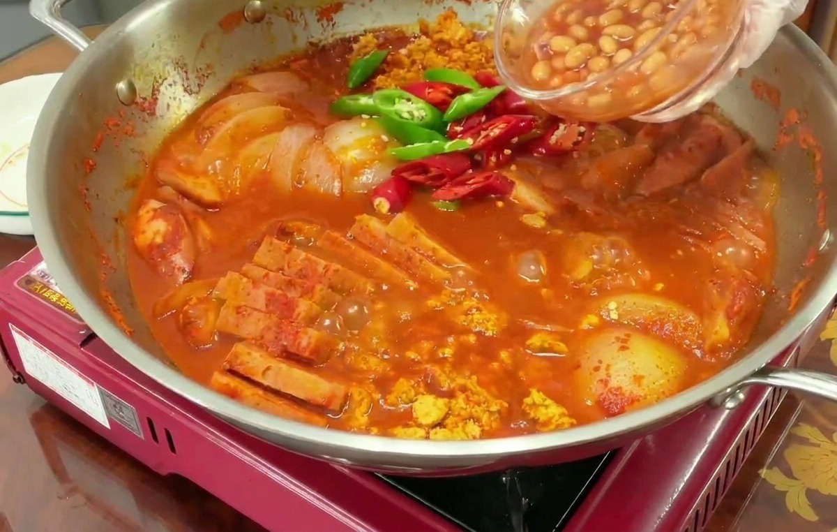 Dłoń przesypuje fasolkę z puszki z małej, szklanej miseczki do bulgoczącego, czerwonego gulaszu z dodatkiem zielonych i czerwonych papryczek chili.