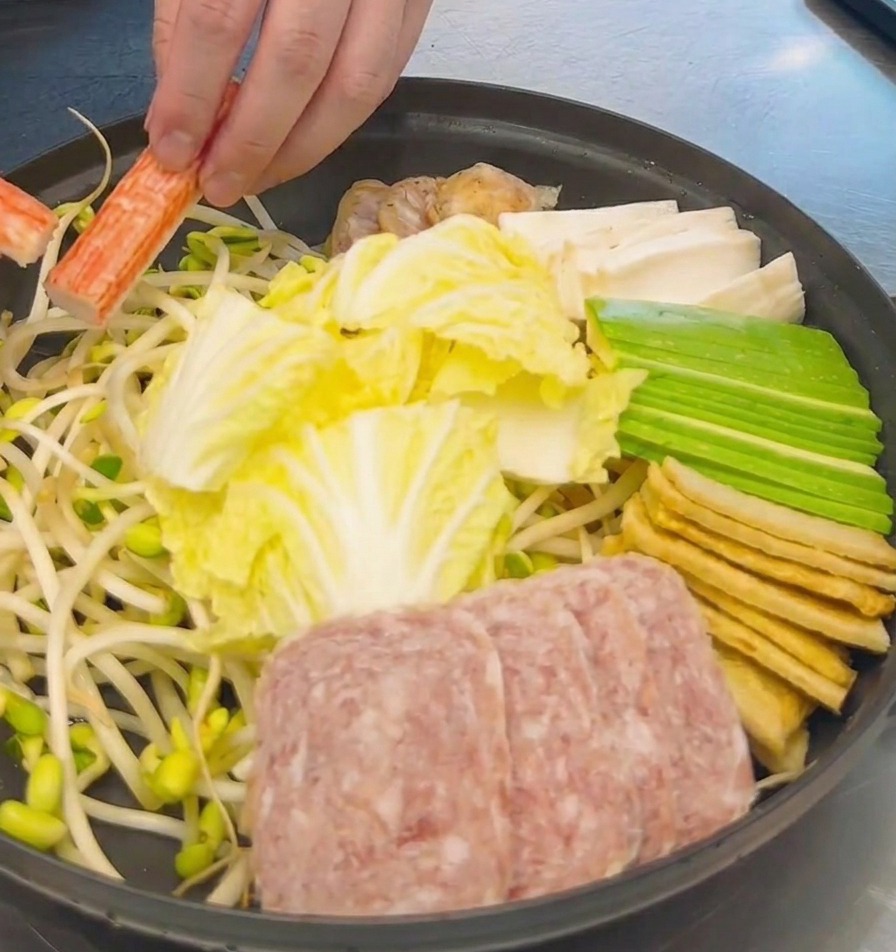 Dłoń układająca pomarańczowo-białe paluszki krabowe w garnku z kapustą, mielonką i tofu.