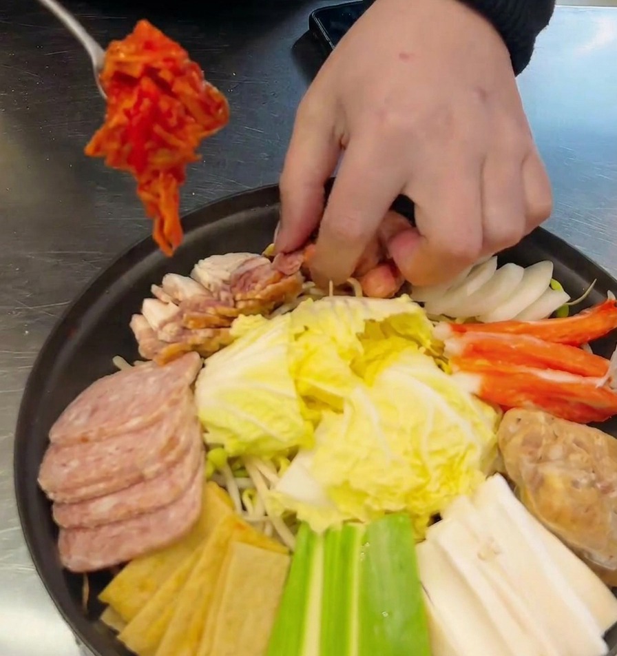 Dłoń nakładająca łyżką porcję czerwonego kimchi do garnka z parówkami i mielonką.