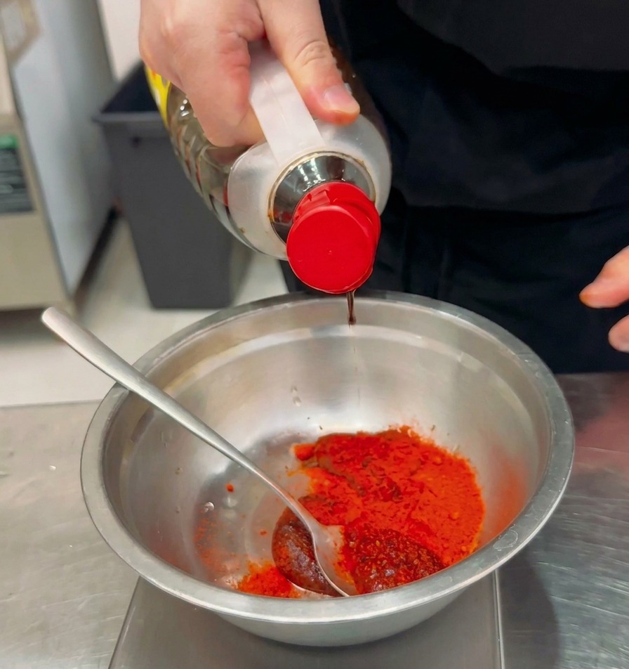 Płynna przyprawa wlewana z butelki do miski z płatkami chili i pastą gochujang.