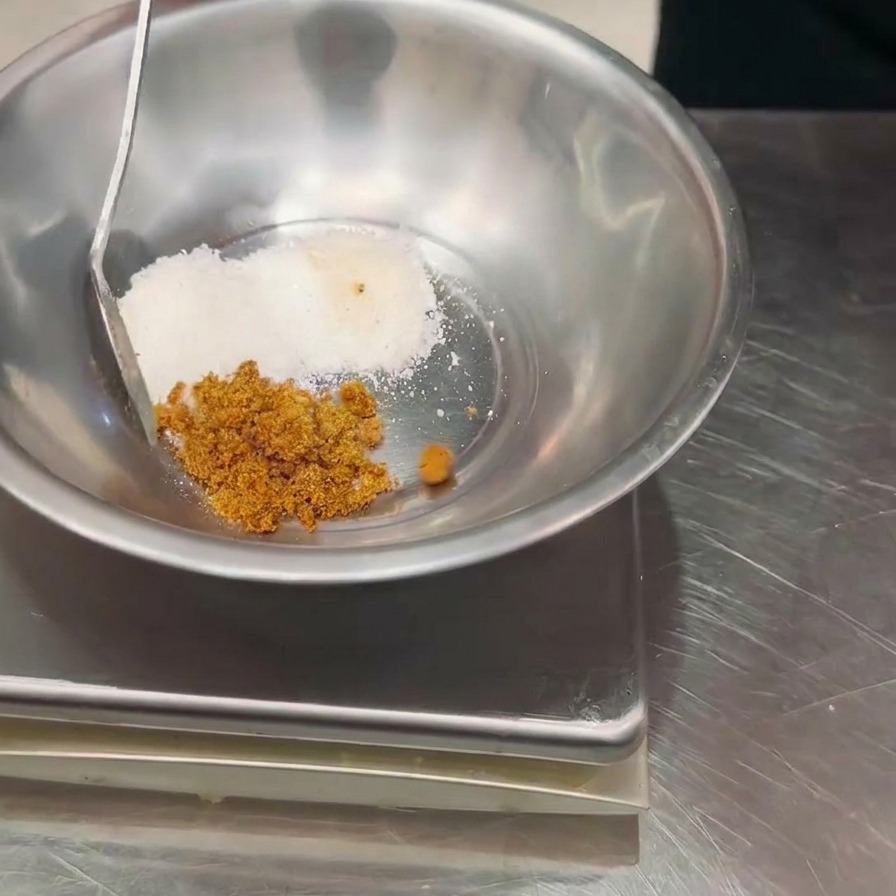 Dłoń dodająca łyżką brązową przyprawę do miski z białym proszkiem na wadze kuchennej.