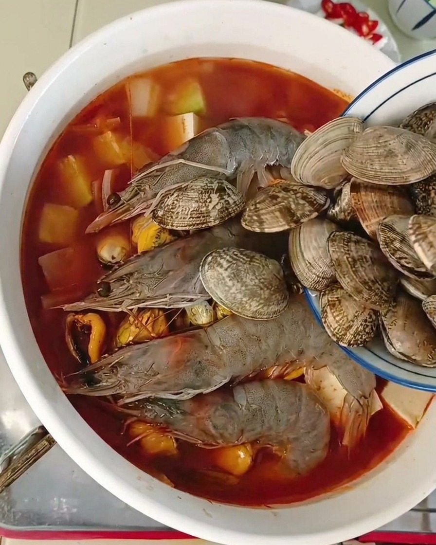 Wrzucanie świeżych małży z niebieskiej miseczki do garnka z krewetkami i tofu w pikantnym bulionie.