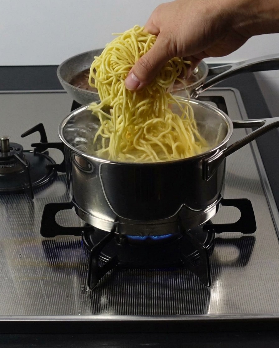 Dłoń wkładająca porcję grubego, żółtego makaronu ramen do garnka z wrzącą wodą.