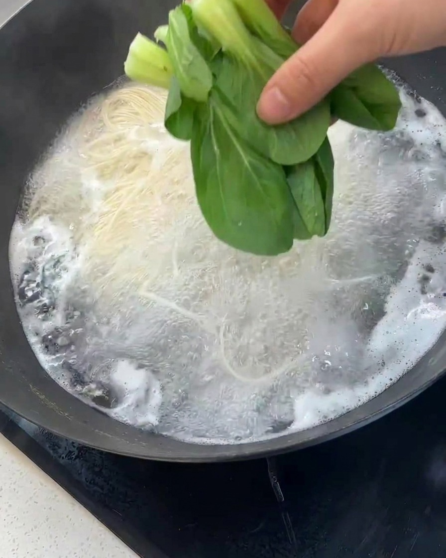 Ręka wrzucająca świeżą kapustę bok choy do woka z gotującym się makaronem.