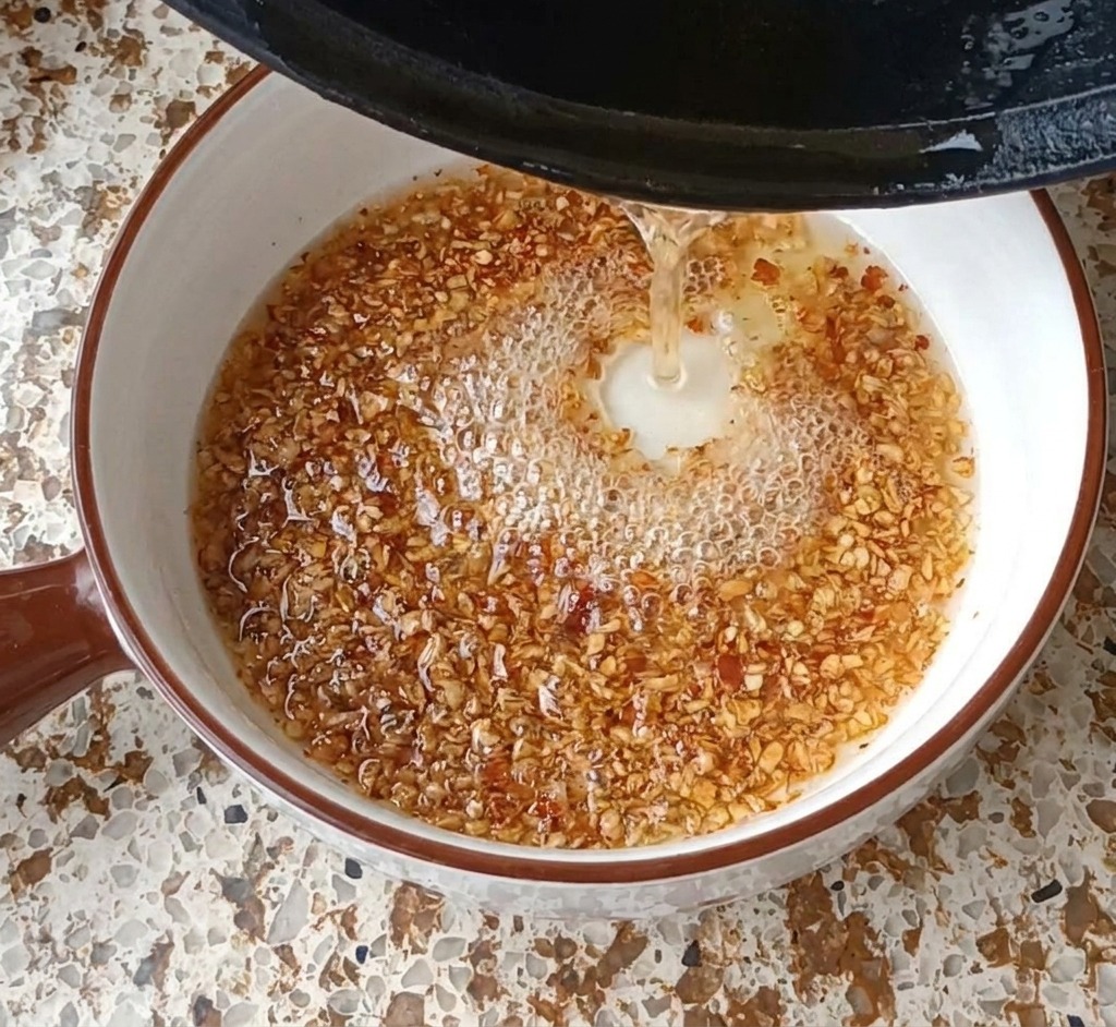 Gorący olej wlewany z czarnej patelni do białej miski z czosnkiem i chili, tworzący bąbelki.