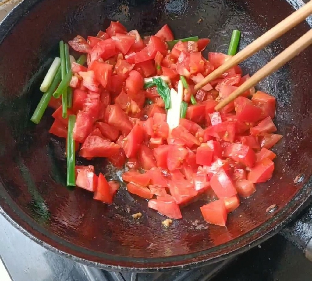 Pokrojone pomidory i dymka smażone w ciemnym woku przy użyciu drewnianych pałeczek.