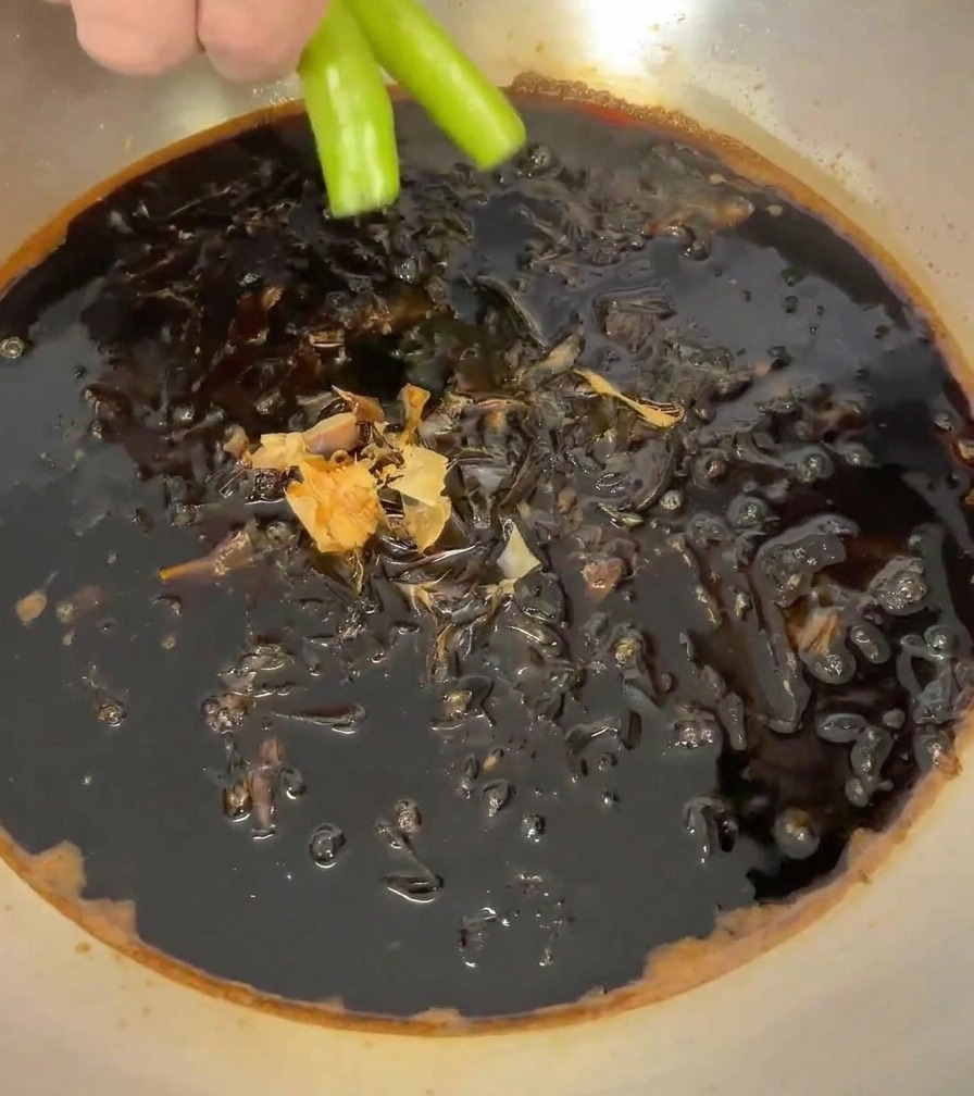 Dłoń kładąca dwie jaskrawozielone papryczki chili do garnka wypełnionego sosem i płatkami bonito.