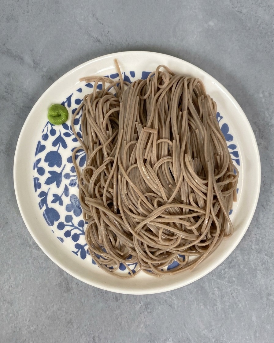 Porcja ugotowanego makaronu soba na biało-niebieskim talerzu z małą porcją zielonej pasty wasabi obok.