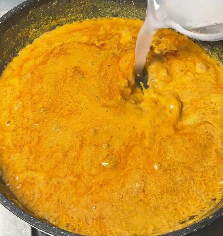 Przezroczysty płyn wlewany z małej miseczki do gotującego się, gęstego, pomarańczowego sosu curry.