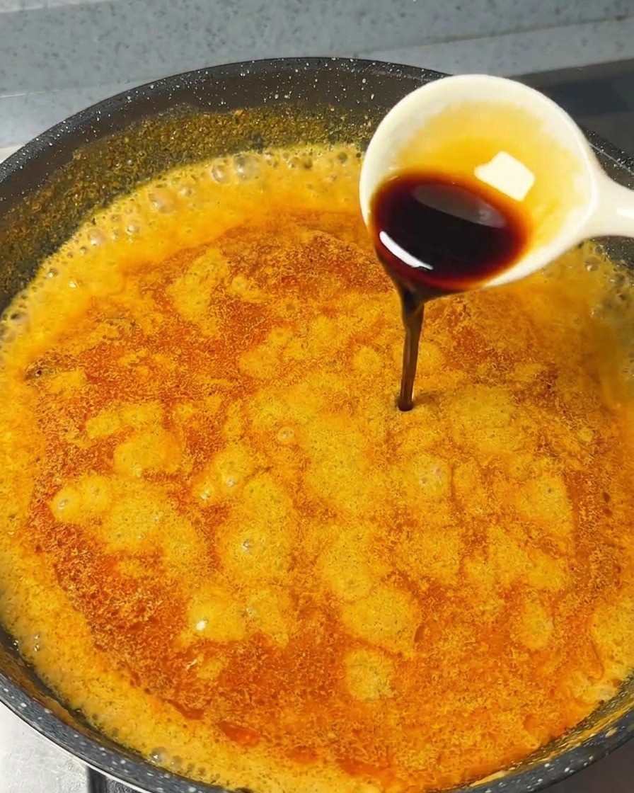 Biała łyżka wlewająca ciemny sos sojowy do gotującego się pomarańczowego sosu curry na patelni.