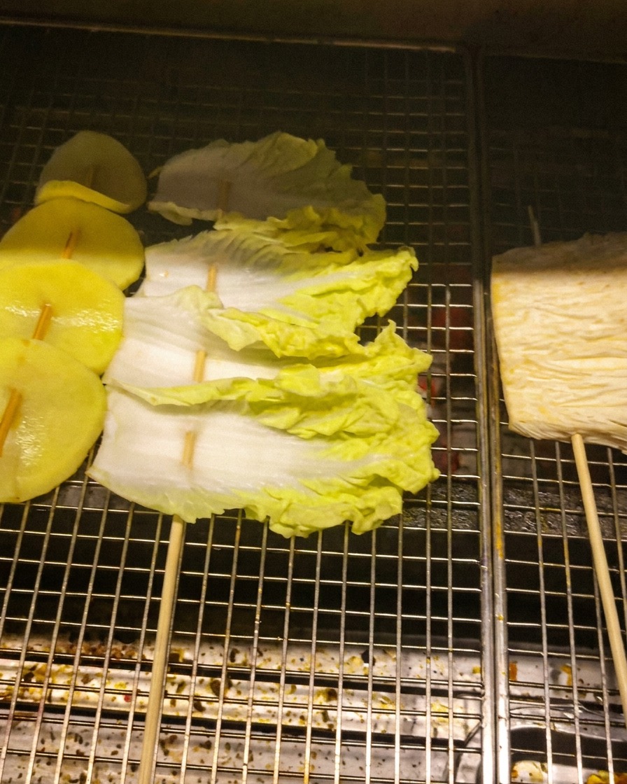 Plastry ziemniaków, liście kapusty i grzyby enoki nadziane na patyczki na metalowym ruszcie grillowym.