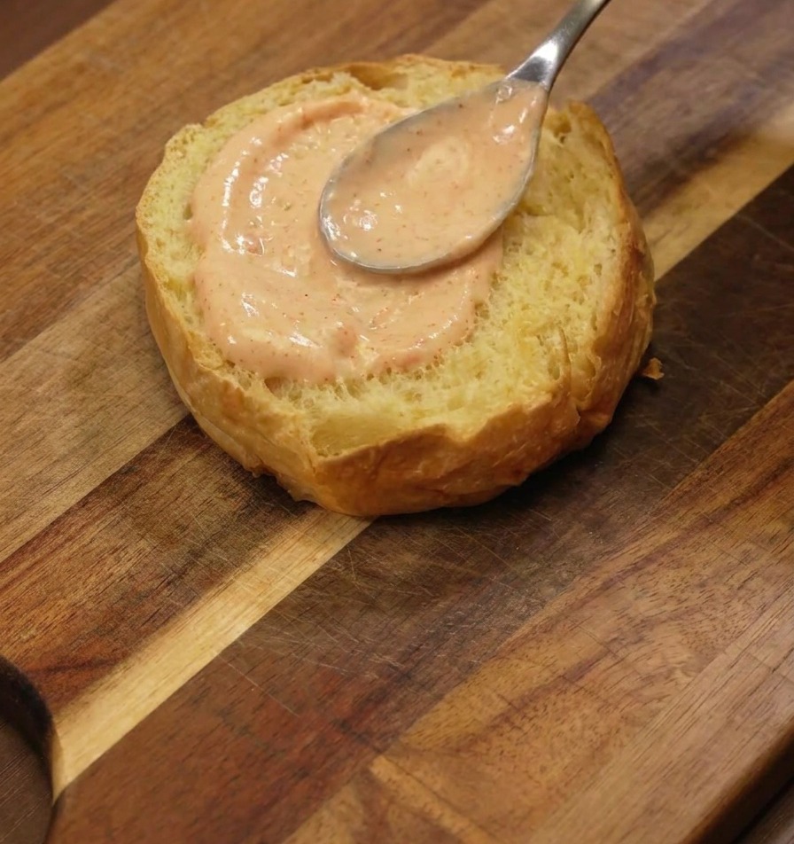 Srebrna łyżka nakładająca grubą warstwę pomarańczowego sosu na opieczoną stronę bułki brioche na desce.