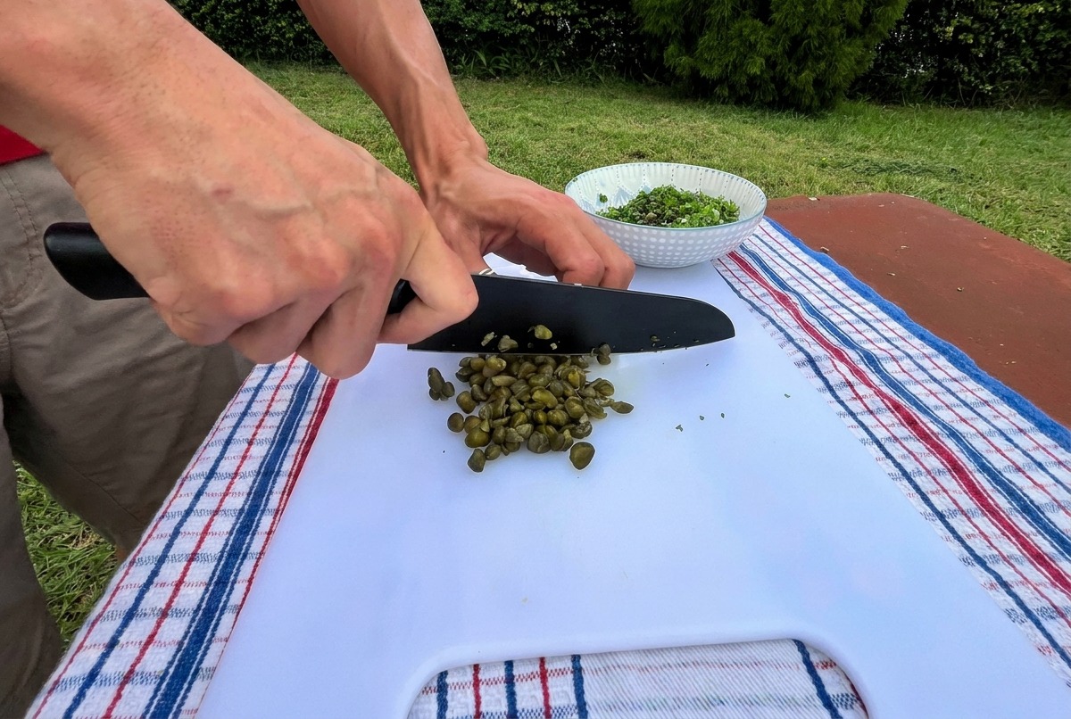 Zbliżenie na siekanie zielonych kaparów czarnym nożem na białej desce na zewnątrz.