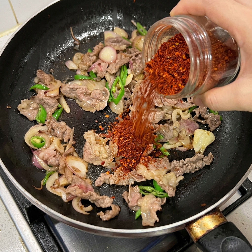 Czerwone chili w proszku sypane z słoiczka na wołowinę, cebulę i zielone papryczki na patelni.