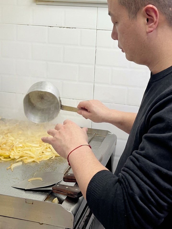Szef kuchni wylewający wodę z małej metalowej chochli na stos mięknącej cebuli na profesjonalnym grillu.