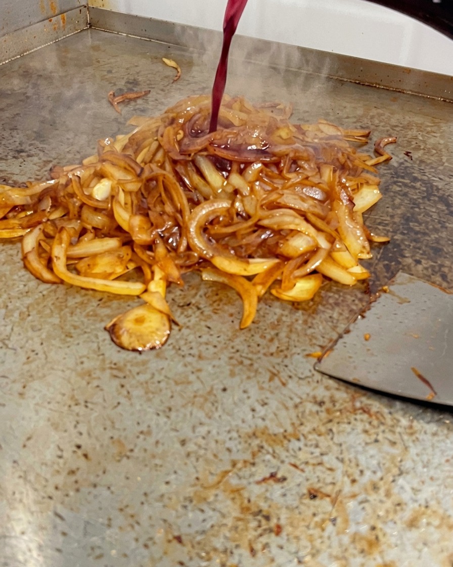Czerwone wino wylewane z butelki na stos mocno zrumienionej cebuli na grillu płaskim.