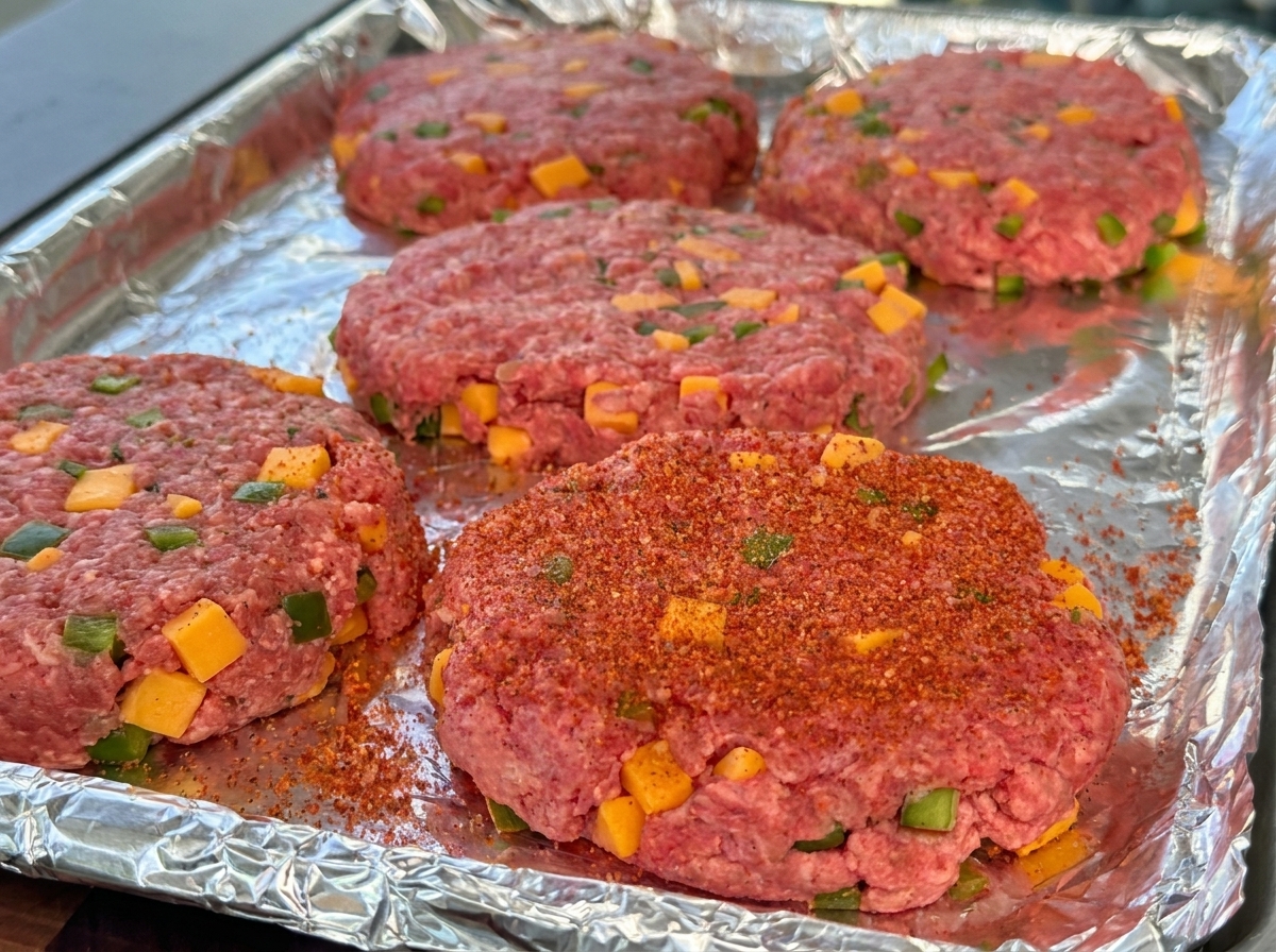 Uformowane surowe kotlety wołowe z widocznymi kostkami sera i zieloną papryką, posypane czerwoną przyprawą BBQ, na tacy z folią aluminiową.