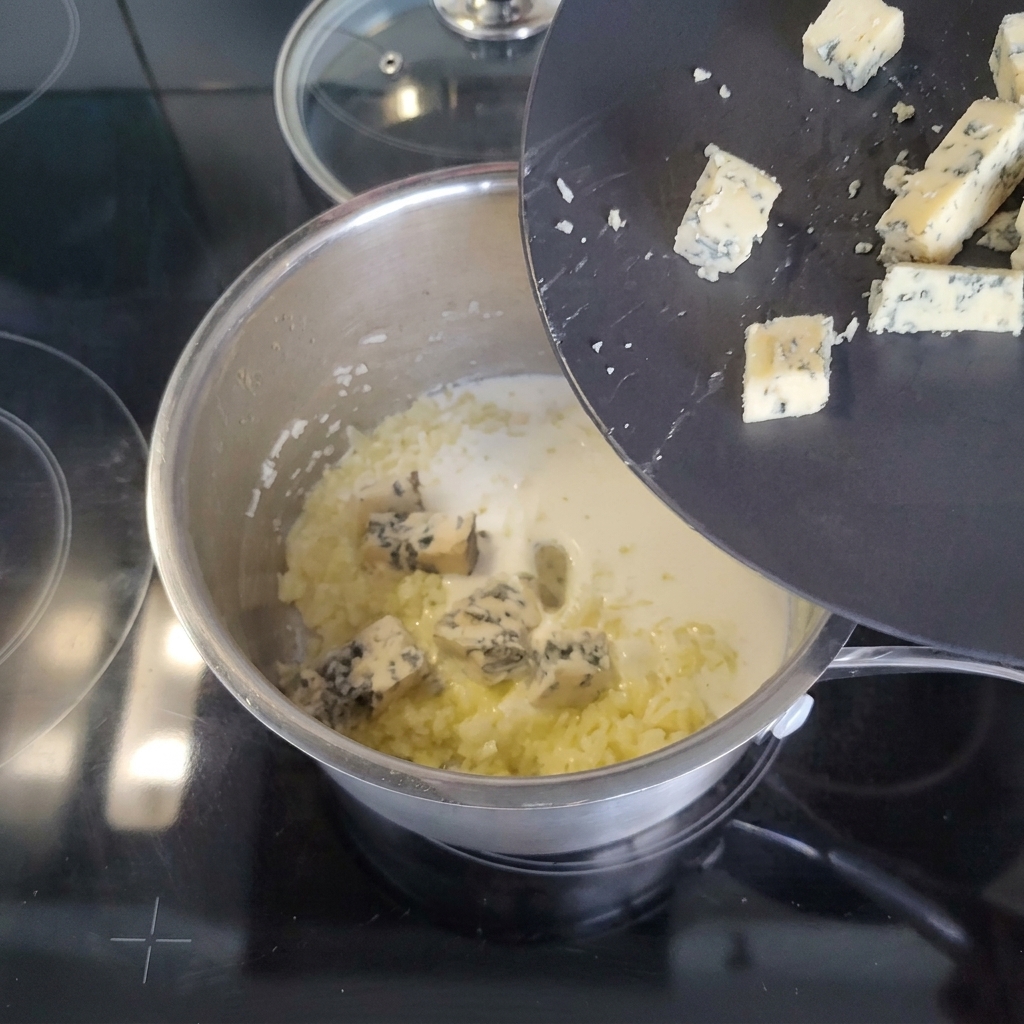 Kawałki sera pleśniowego zsuwające się z nachylonego czarnego talerza do rondelka ze śmietanką i podsmażonymi warzywami.