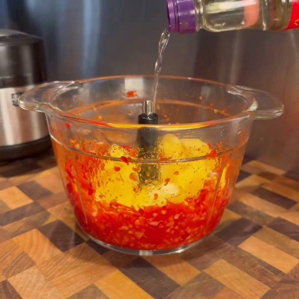 Płyn wlewany z butelki do szklanej misy blendera z czerwoną pastą chili i żółtymi kawałkami mango.