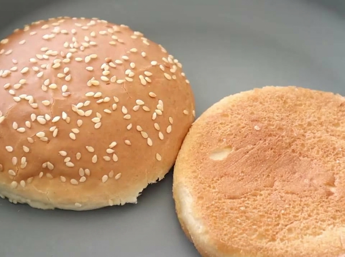 Dwie połówki bułki burgerowej z sezamem leżące obok siebie, z których jedna jest idealnie złocisto-brązowa.
