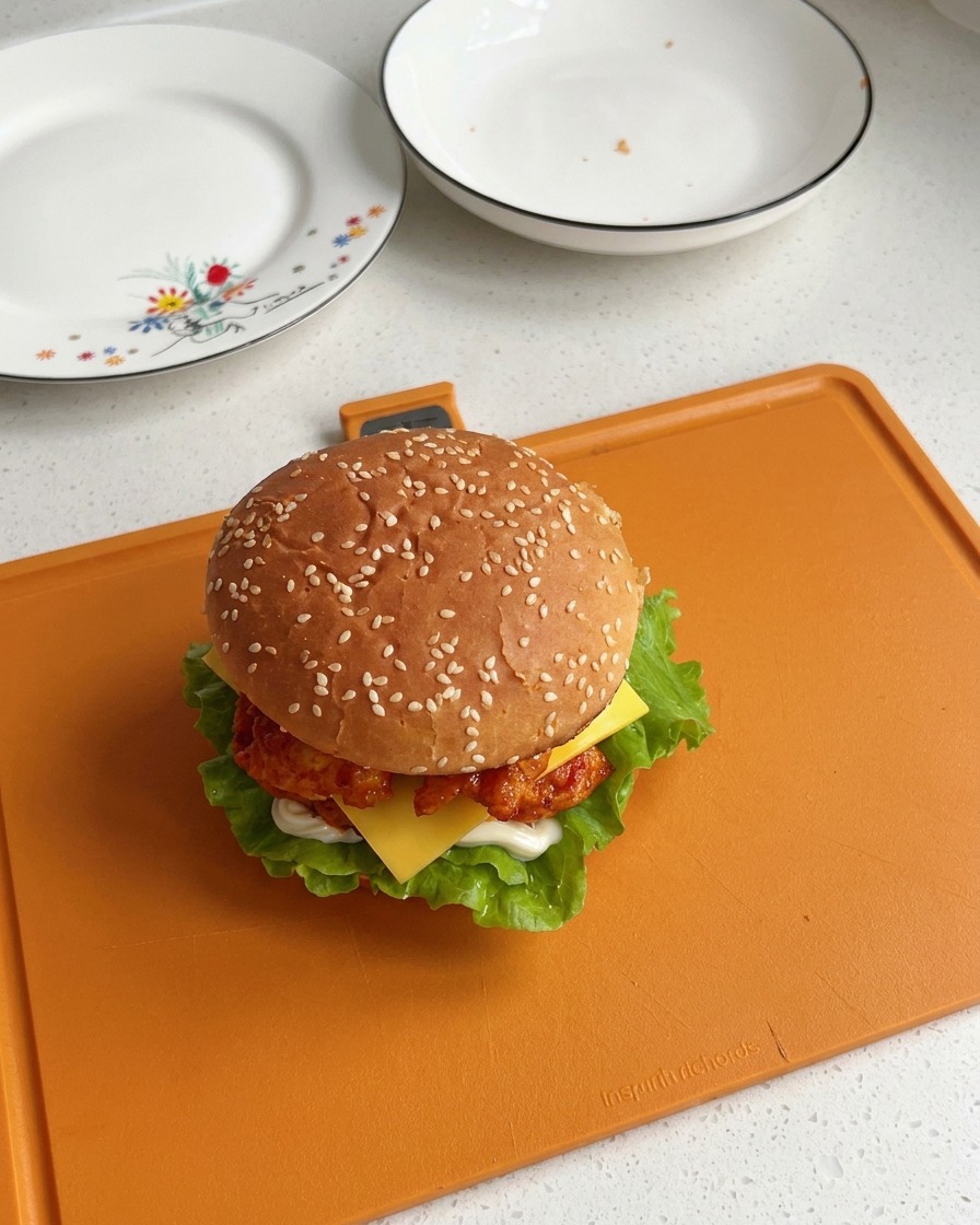 W pełni złożony burger z kurczakiem z Orleanu przykryty bułką z sezamem, pokazujący widoczne warstwy sałaty, kurczaka i topiącego się plastra sera.