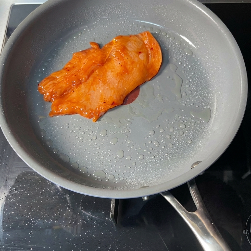 Zamarynowany, pomarańczowy kotlet z kurczaka smażący się na szarej patelni z widocznymi kropelkami oleju.