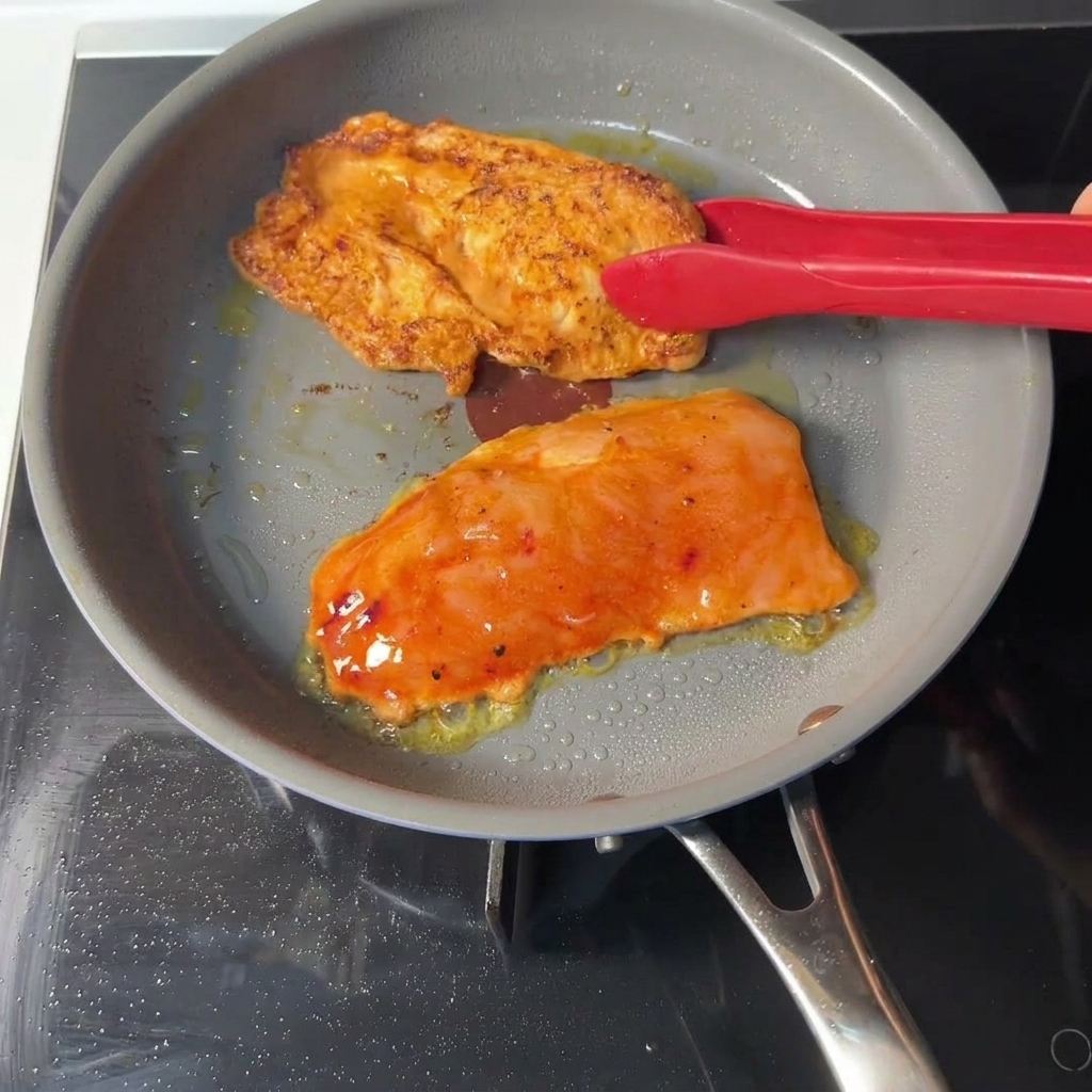 Czerwone szczypce kuchenne obracające pomarańczowy kotlet z kurczaka na patelni, odsłaniając rumianą, dobrze wysmażoną skórkę.