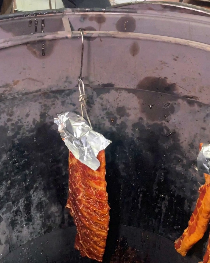 Płat zamarynowanych żeberek wieprzowych z folią aluminiową chroniącą górną część, wiszący wewnątrz głębokiego, ciemnego cylindrycznego pieca do pieczenia.