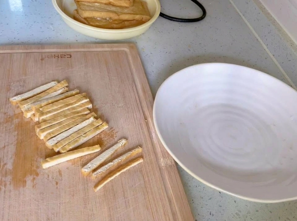 Złocistobrązowe usmażone tofu pokrojone w równe, cienkie paski na drewnianej desce obok białego talerza.