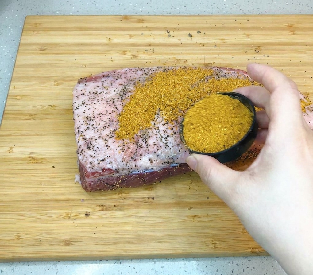 Dłoń trzymająca małą czarną miseczkę i posypująca złocistobrązową przyprawą barbecue duży kawałek surowego mostka wołowego na drewnianej desce.