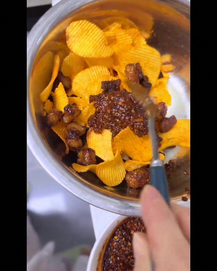 Dłoń za pomocą łyżki dokładnie miesza karbowane chipsy, chrupiący tłuszcz wołowy i krwistoczerwony olej chili w metalowej misce.