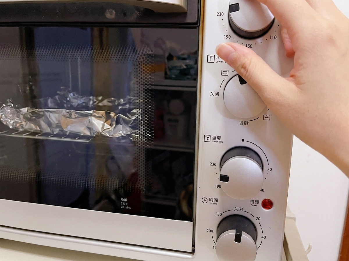 Zbliżenie na dłoń ustawiającą pokrętło temperatury w białym elektrycznym piekarniku na 230°C, w środku widoczne zawinięte w folię produkty.