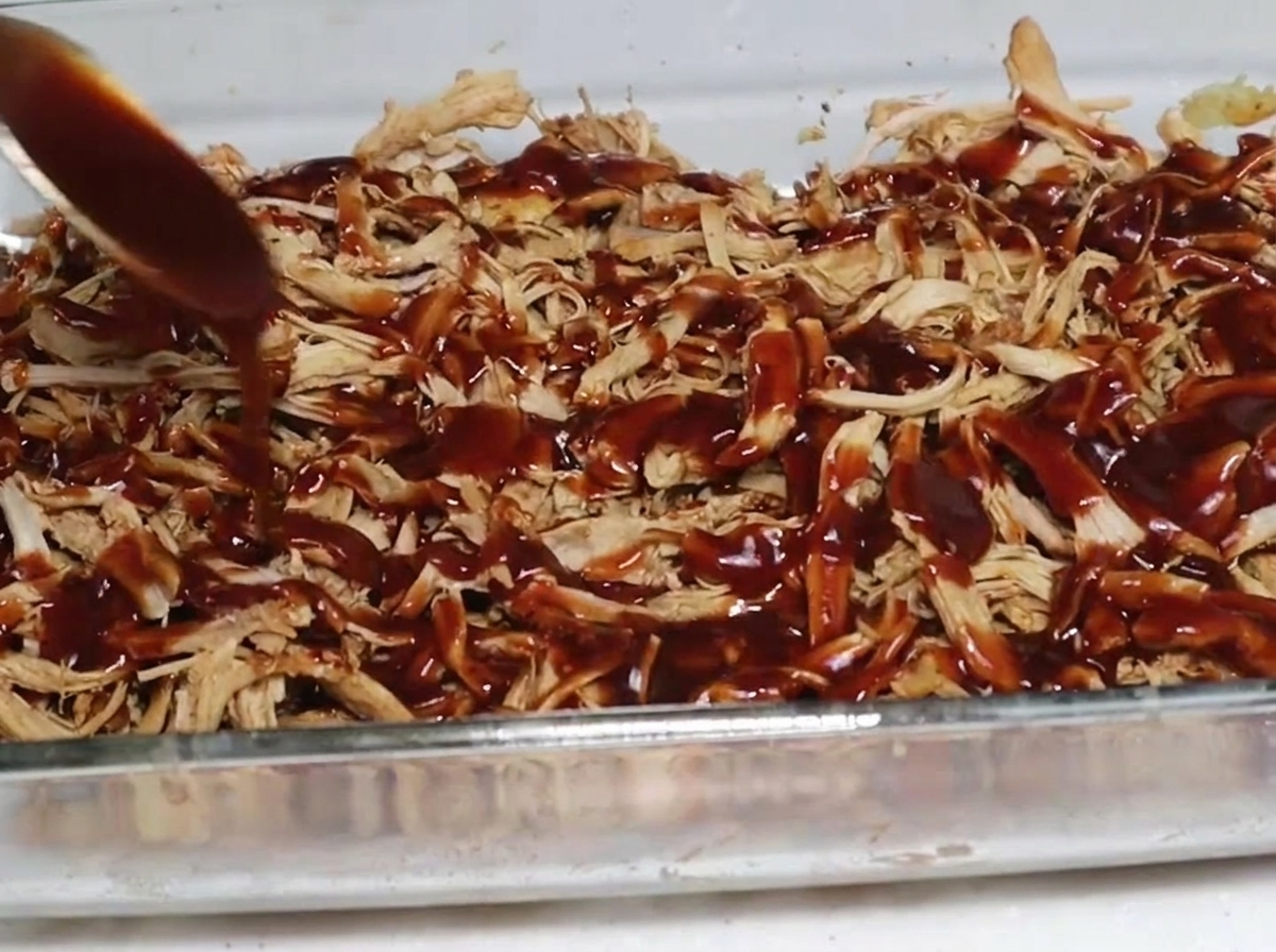 Łyżka z gęstym, ciemnym sosem barbecue polewająca wędzoną, szarpaną wieprzowinę w szklanym naczyniu.