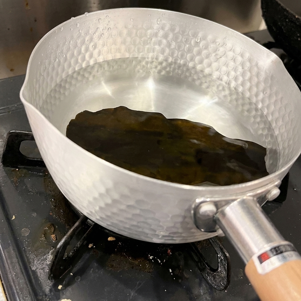 Pojedynczy kawałek ciemnego, suszonego wodorostu kombu spoczywa na dnie aluminiowego garnka wypełnionego czystą, zimną wodą.