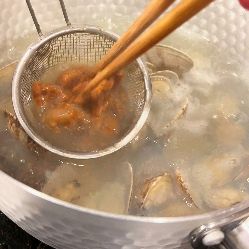 Małe podręczne sitko trzyma brązową pastę miso, podczas gdy pałeczki delikatnie rozprowadzają ją w gorącej zupie z małżami.