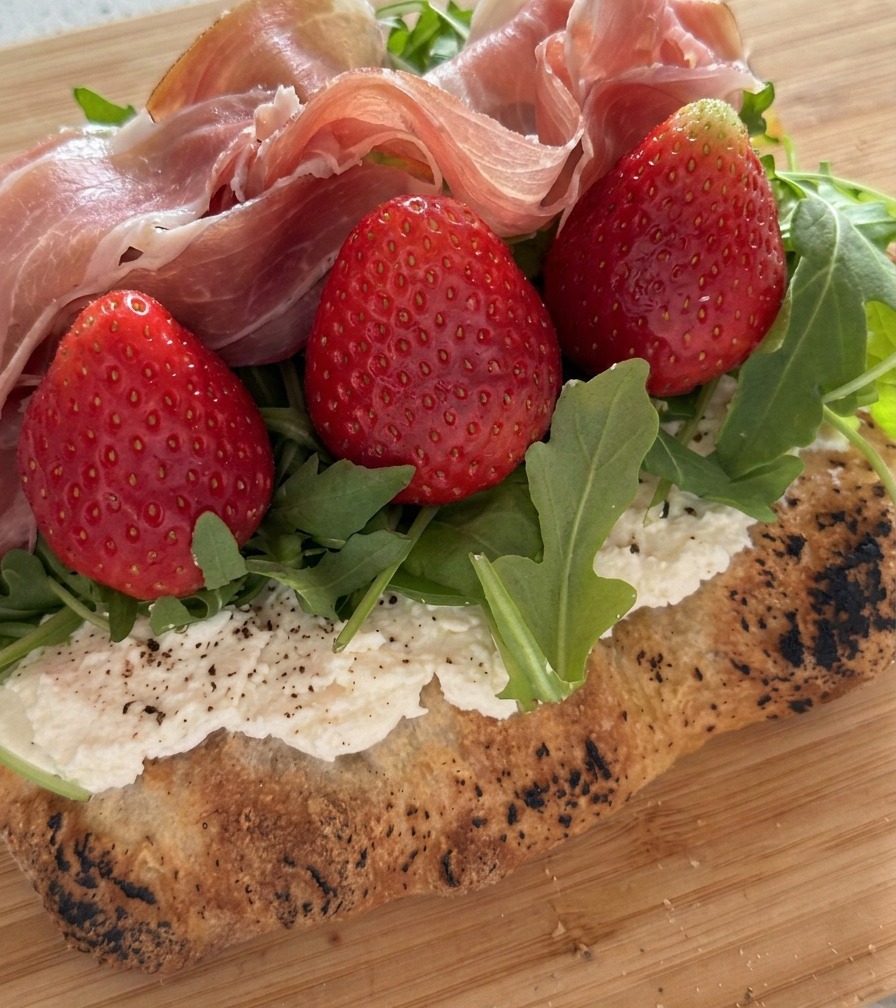 Zbliżenie na złożone plasterki prosciutto układane na świeżych truskawkach i rukoli.