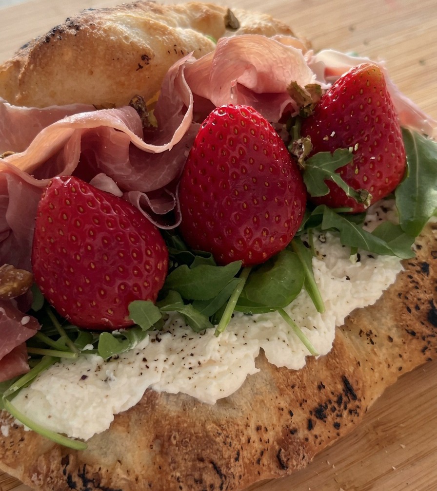 Pokruszone pistacje posypywane na kanapkę wypełnioną prosciutto, truskawkami i rukolą.
