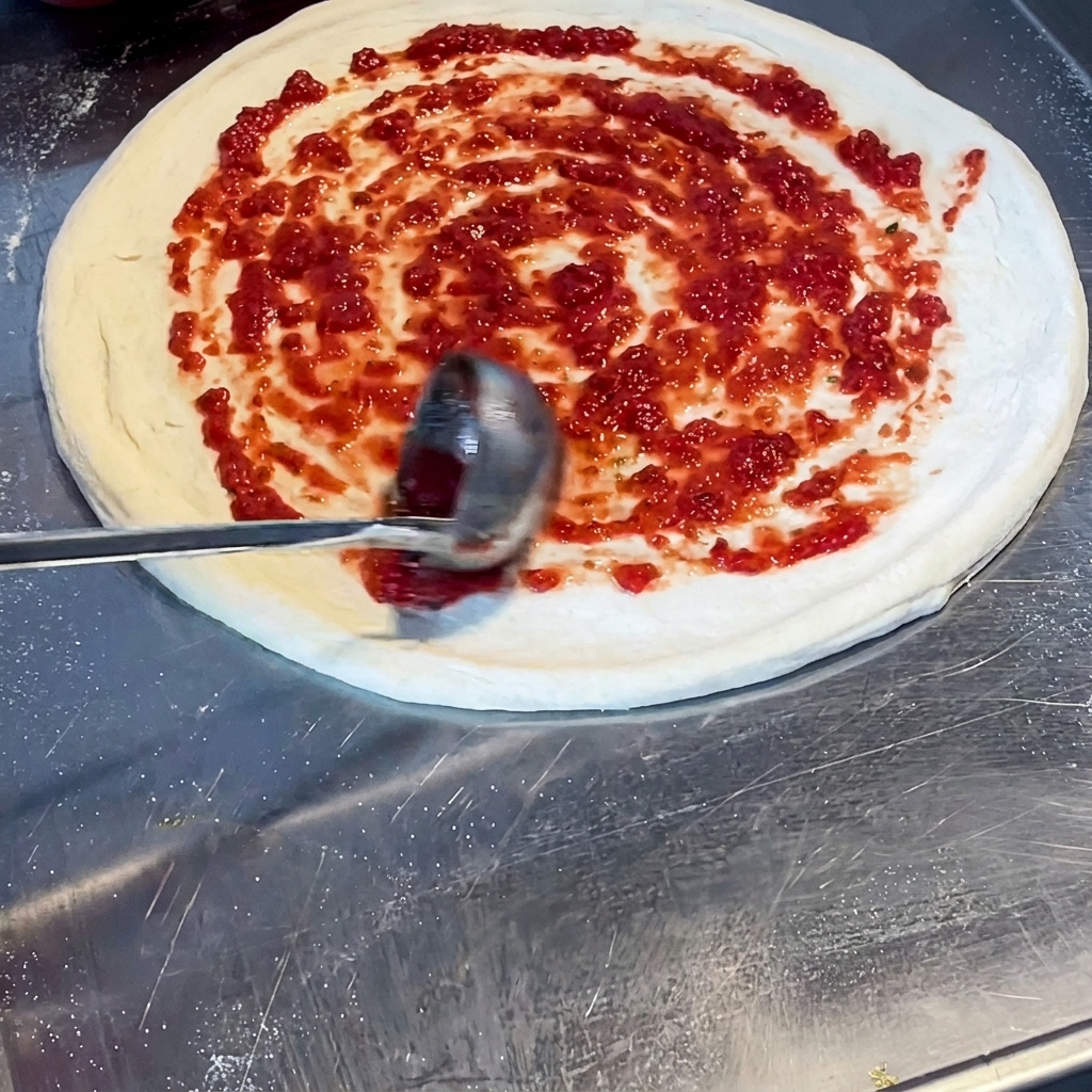 Chochla rozprowadzająca jaskrawoczerwony sos pomidorowy w spiralny wzór na surowym cieście pizzy.