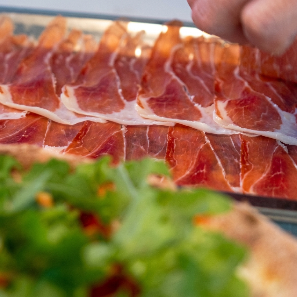 Cienkie plastry szynki prosciutto ostrożnie układane na pizzy z rukolą.