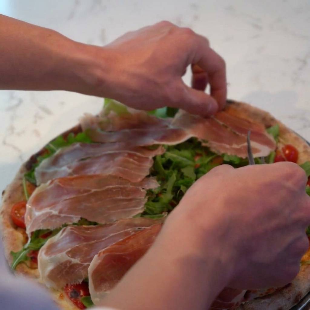 Starty ser parmezan posypywany na pizzę, pokrywający prosciutto i rukolę.