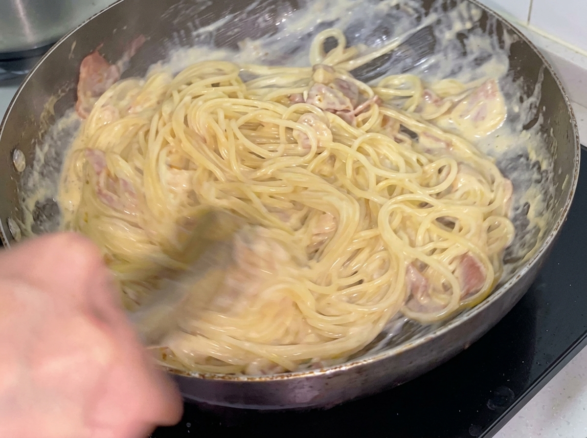 Kremowe spaghetti mieszane z boczkiem i kurczakiem za pomocą metalowych szczypiec na patelni.