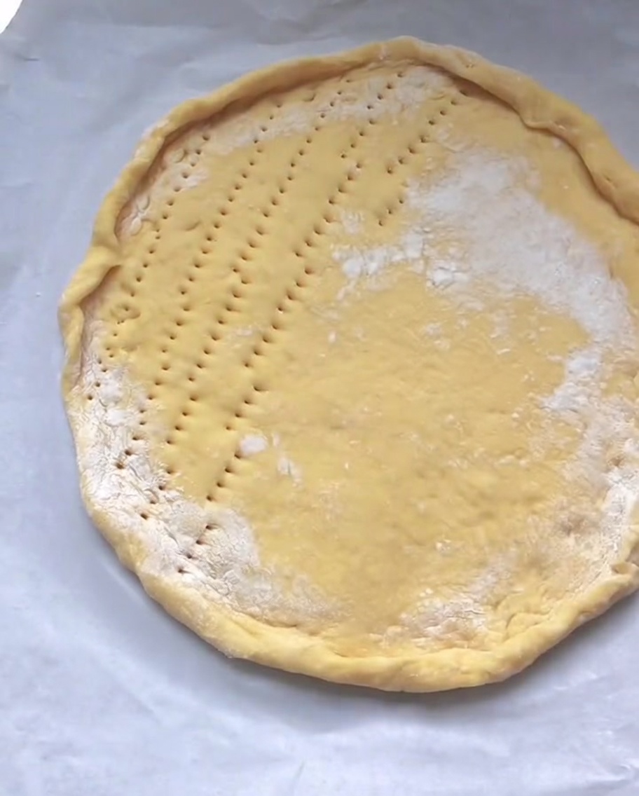 Surowy okrągły spód pizzy na papierze do pieczenia z równomiernie zrobionymi dziurkami na płaskiej powierzchni.