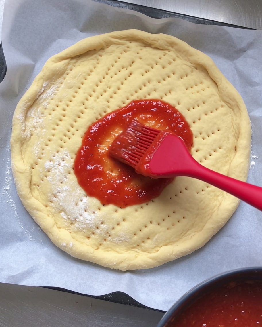Czerwony sos pomidorowy równomiernie rozsmarowywany na surowym, nakłutym spodzie pizzy.