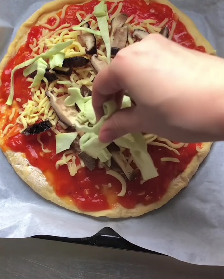 Dłoń nakładająca poszatkowaną zieloną kapustę na pizzę pokrytą sosem pomidorowym, serem i pokrojonymi grzybami.