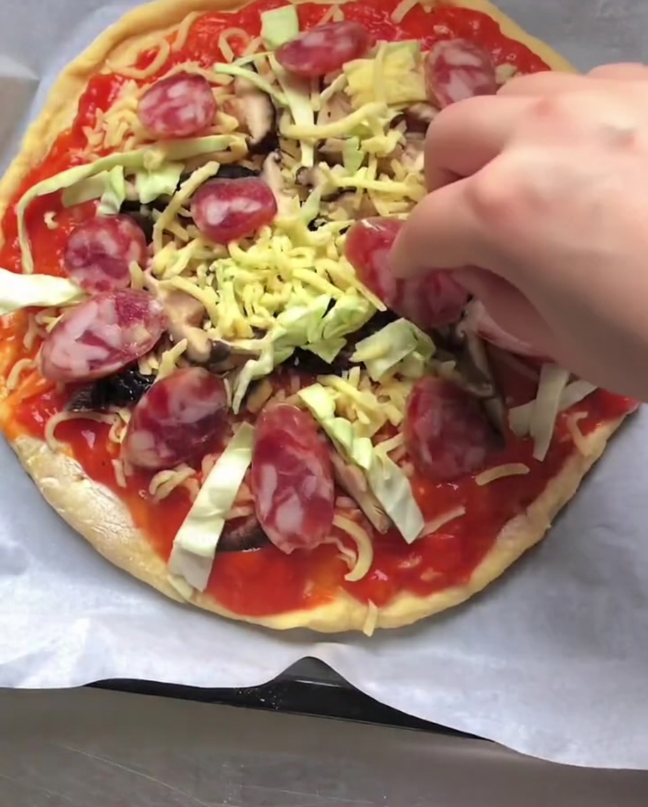 Dłoń układająca plasterki czerwonej włoskiej kiełbasy na surowej pizzy pokrytej sosem, serem i warzywami.