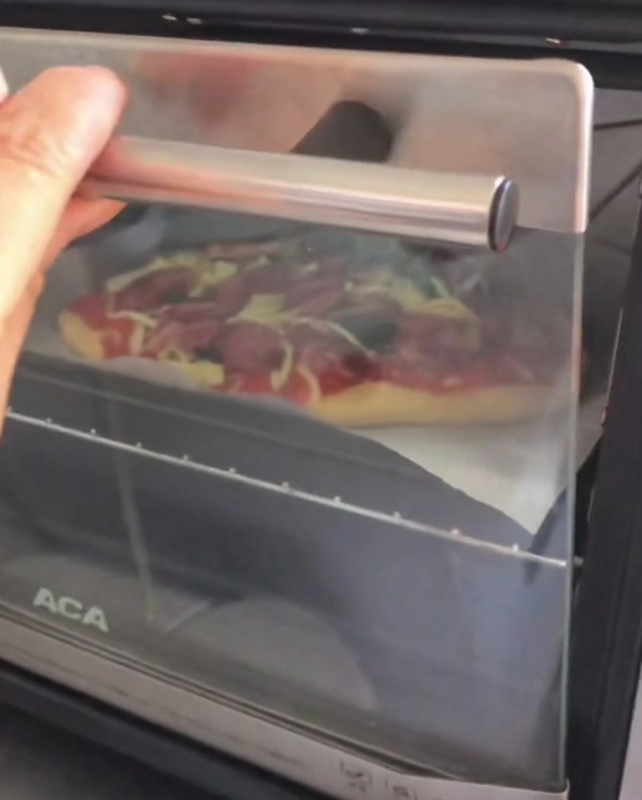 Otwieranie drzwiczek małego piekarnika nablatowego, aby włożyć do niego gotową, surową pizzę na blaszce.