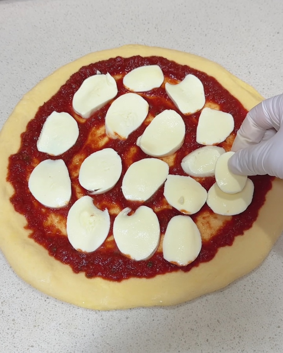 Dłoń w rękawiczce kładąca grube plastry świeżej mozzarelli na spód do pizzy pokryty czerwonym sosem pomidorowym.