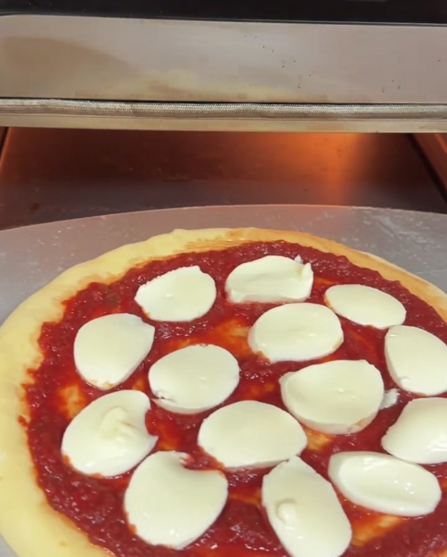 Surowa pizza z sosem i mozzarellą wsuwana do elektrycznego pieca do pizzy na metalowej łopacie.