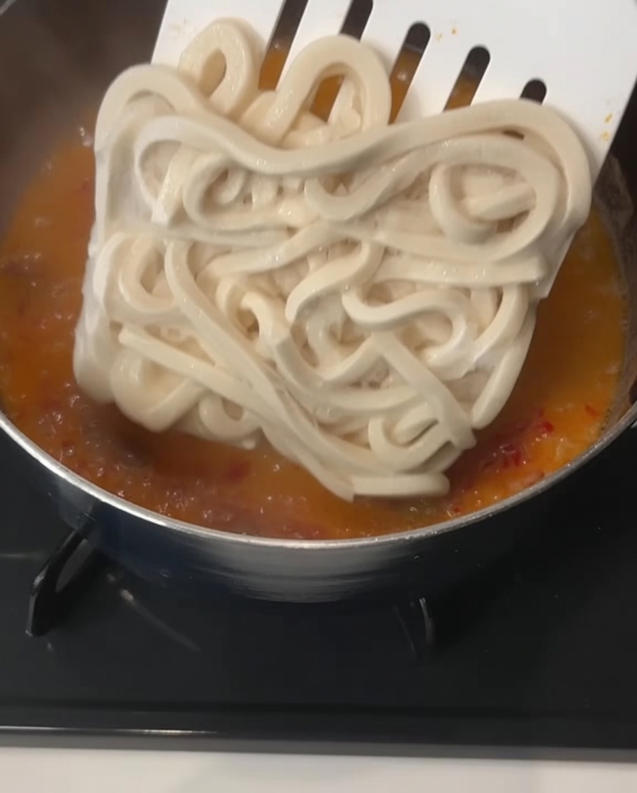 Duża, kwadratowa kostka grubego mrożonego makaronu udon wkładana szpatułką do gotującego się, kremowego pomarańczowego wywaru.